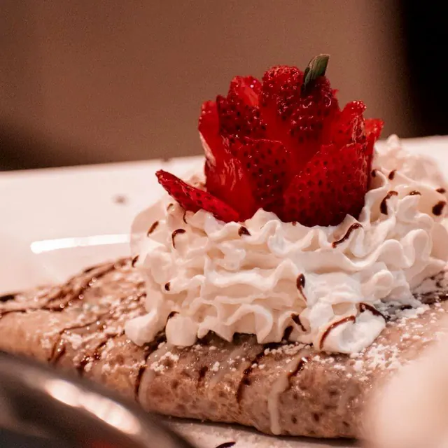 A folded crepe topped with ruffled whipped cream, a strawberry cut into a rose, chocolate drizzle and powdered sugar