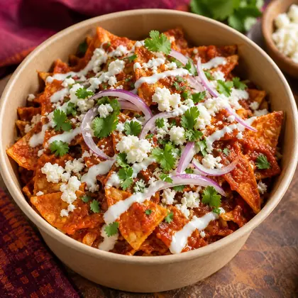 A bowl of red salsa chilaquiles topped with crema, cheese, onion and cilantro