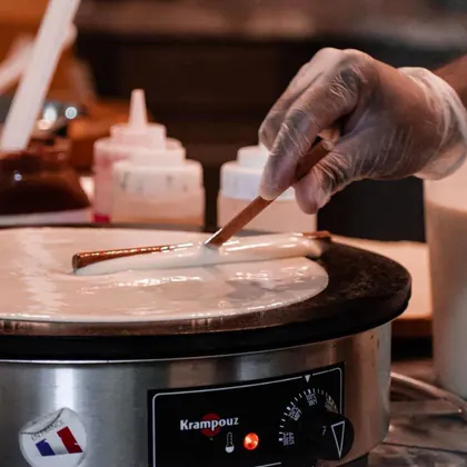 A gloved hand spreading crepe batter thin with a wooden spreader across a round Krampouz griddle, with squeeze bottles behind