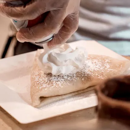 A gloved hand topping a folded crepe with whipped cream, over a dusting of powdered sugar on a square white plate