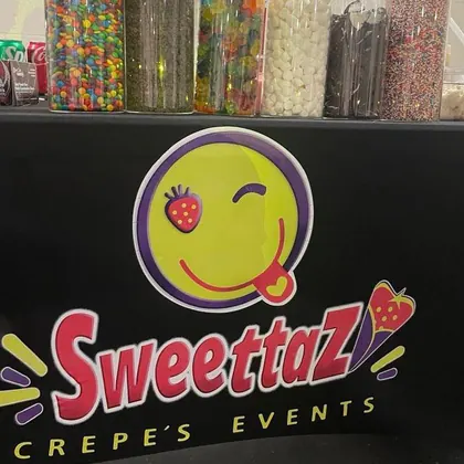Tall jars of M&M's, chocolate chips, gummy bears, marshmallows, Oreo cookies and rainbow sprinkles on a black table printed with the Sweettaz Crepe's Events logo